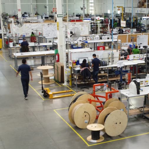 Industrial manufacturing facility with workers assembling and testing products, large wooden cable spools, and organized workstations