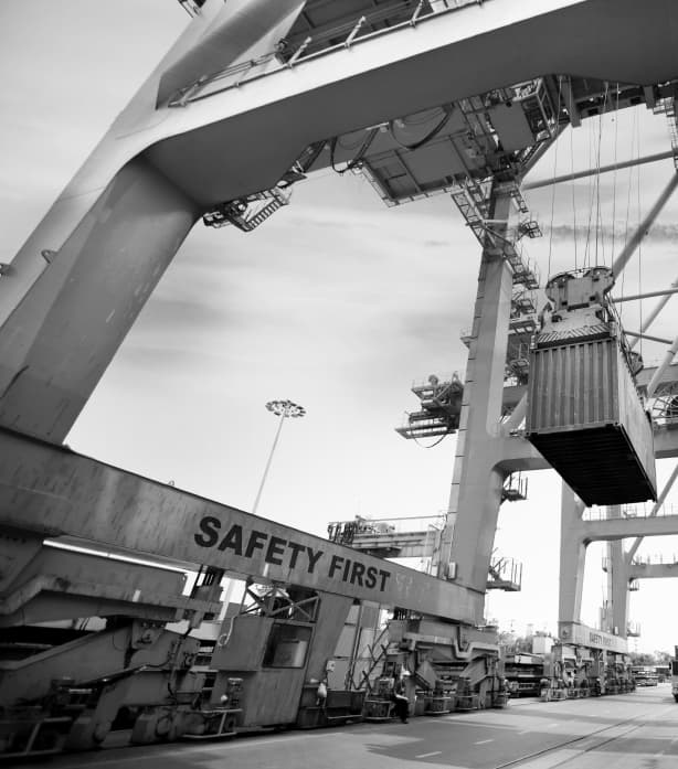 Shipping container being lifted by a large port crane with "SAFETY FIRST" sign visible