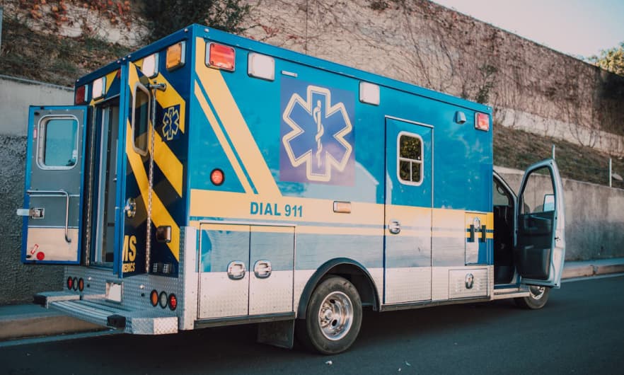 A blue and yellow ambulance with its doors open, parked on the side of a road next to a concrete wall.