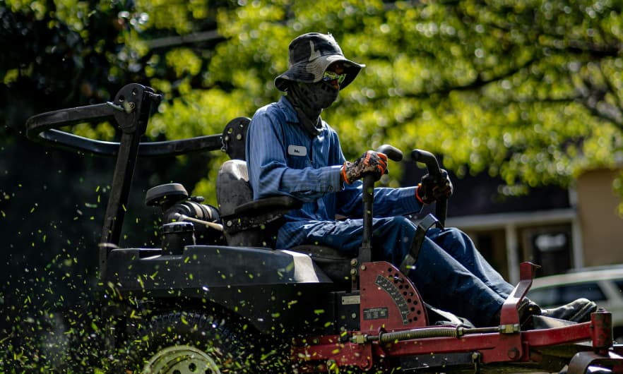A worker wearing protective clothing and a hat operates a riding lawn mower, surrounded by flying grass clippings.